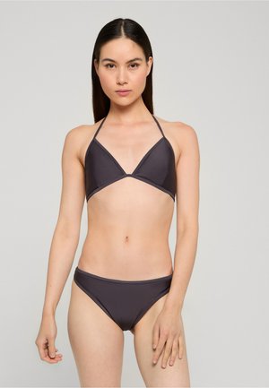 Kobieta o długich ciemnych włosach ubrana w ciemnoszary bikini z wiązaniem na szyi oraz pasujące do niego figi, stoi na tle jednolitego jasnoszarego tła.