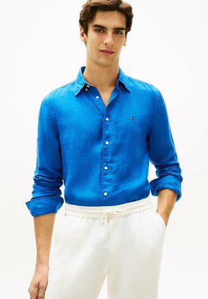 Jonge man die een blauw overhemd met opgerolde mouwen en witte broek met trekkoord draagt, staand met handen in de zakken.