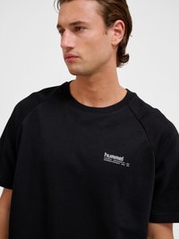 Junger Mann mit kurzen braunen Haaren, der ein schwarzes Hummel-T-Shirt trägt und nach links vor einem schlichten weißen Hintergrund schaut.