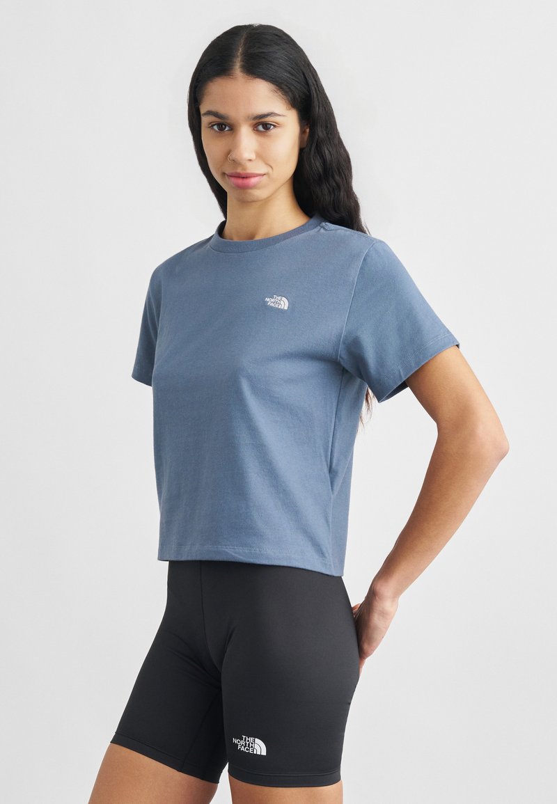 Frau mit langem, dunklem Haar, die ein blaues, bauchfreies T-Shirt und eine schwarze, eng anliegende Shorts trägt, beide mit The North Face-Logos, steht vor einem schlichten Hintergrund.