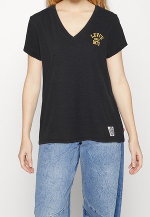 Henkilö, jolla on yllään musta V-pääntiellä varustettu Levi's T-paita, jossa on keltainen logo rinnassa, sekä vaaleansiniset farkut, seisoo käsivarret rentoina sivuilla.