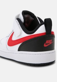 Baskets blanches Nike avec swoosh rouge et détail talon noir, dotées d'une languette noire et d'une fermeture à bande velcro blanche.