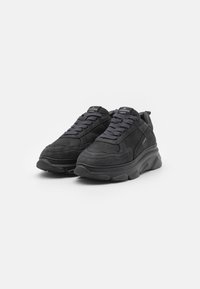 Musta sneakerit, joissa on yhdistelmä mokkaa ja sileää kangasta. Niissä on paksut pohjat, huomattava nauhoitus sekä brändäys kielessä ja kantapäässä.
