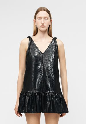 Γυναίκα που φοράει μίνι αμάνικο μαύρο φόρεμα από ψεύτικο δέρμα φιδιού με V-λαιμόκοψη και βολάν στο τελείωμα, στέκεται μπροστά σε απλό γκρι φόντο.