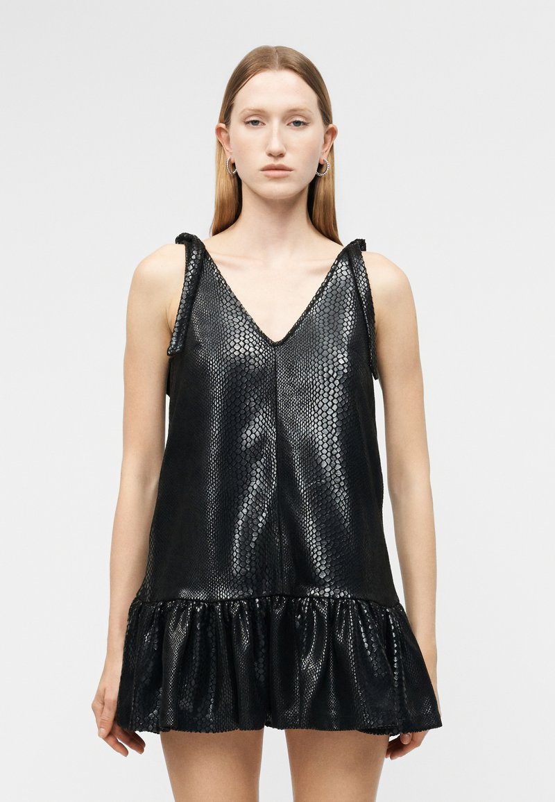 Kobieta w czarnej mini sukience bez rękawów z imitacji skóry węża, z dekoltem w serek i falbaną na dole, stojąca na jednolitym szarym tle.