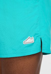 Pantaloni da bagno turchesi realizzati in tessuto leggero, con orlo cucito e una toppa grafica di una sneaker bianca sulla gamba inferiore.