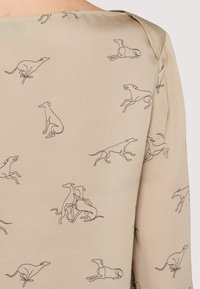 Beżowy materiał z wzorem w postaci rysunków linii owczarków w różnych pozach. Gładka tekstura, luźny krój i okrągły dekolt.