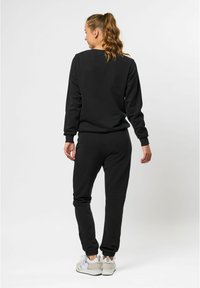 Svart träningsbyxor och matchande crewneck sweatshirt i mjukt tyg. Båda har ribbade muddar och en avslappnad passform. Bärs med vita träningsskor.