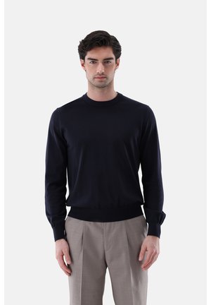 Mann mit dunklem Haar, der einen schwarzen Pullover mit langen Ärmeln und hellgraue, plissierte Hosen trägt, steht vor einem schlichten weißen Hintergrund.