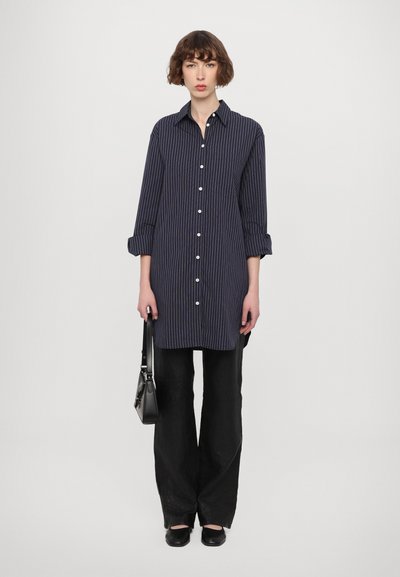 Vestido de camisa a rayas marinas con cuello, cierre de botones y bolsillos. Combinado con pantalones anchos negros y zapatos planos negros, sosteniendo un bolso pequeño.