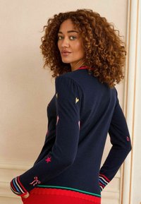 Una donna dai capelli ricci si gira leggermente, mentre indossa un maglione blu navy decorato con ricami colorati di cuori e stelle, su uno sfondo chiaro.