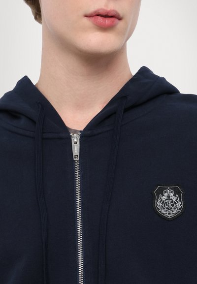 Tumed zip-up kapuutsiga pusa, millel on hõbedane tõmblukk ja vasakul rinnal keerukate mustritega plaastriga. Pehme kangas, millel on tekstureeritud viimistlus.