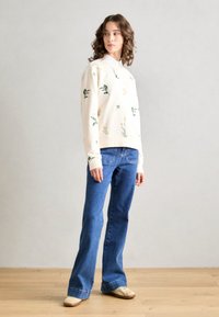 Kremfarget genser med blomsterdesign i grønt og gult, kombinert med blå vide jeans og beige sko. Glatt tekstur, avslappet passform.