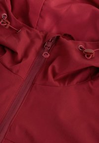 Veste imperméable rouge avec un col haut, des cordons de serrage réglables et une fermeture éclair centrale. La texture du tissu semble lisse et durable.