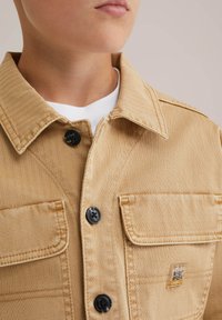 Otrok, oblečen v svetlo rjavo jakno z gumbi, ki ima dve sprednji žep in majhno oznako "Blue Ridge" na desnem žepu, preko bele srajce.