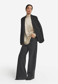 Svart skräddarsydd kavaj över en beige polotröja, kombinerad med vida mörk denimbyxor och stängda klackskor. Modellen står med handen i fickan.