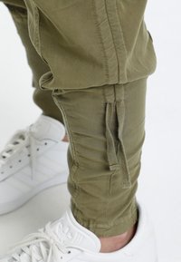 Khaki cargo pants with elastic cuffs, featuring rope drawstrings and side stitching details, paired with white athletic shoes.