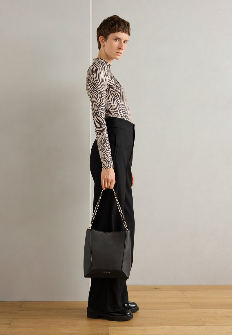 Borsa a secchiello in pelle nera con manico a catena, caratterizzata da una texture liscia e una forma strutturata. Indossata da una persona in una maglietta a strisce zebrate e pantaloni sartoriali.