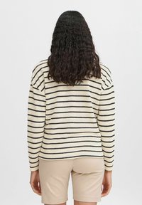 Cremefarbener Pullover mit schwarzen Streifen, strukturierter Stoff, lockere Passform, überschnittene Schultern und lange Ärmel. Getragen mit beigen Shorts.
