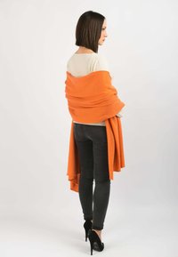 Orange indpakningsjakke draperet over en cremet top, parret med sorte skinny jeans og sorte hæle. Blødt stof, glat tekstur og flydende design.