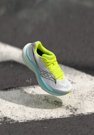 Weißer und neon-gelber Laufschuh mit hellblauer Sohle, der über Asphalt mit weißen Markierungslinien schwebt.