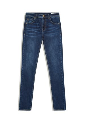 Femme aux longs cheveux blonds portant un débardeur blanc avec un texte rouge, un jean bleu, des sandales noires à talons, tenant un sac à main noir sur fond gris.