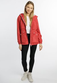 Rote leichte Jacke mit Kapuze, Kordelzug in der Taille und zwei Fronttaschen. Getragen über einem weißen Oberteil und kombiniert mit schwarzen Hosen und weißen Turnschuhen.