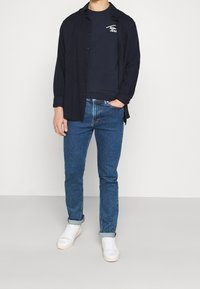 Uomo con camicia aperta blu scuro, t-shirt con logo navy, jeans blu arrotolati e sneaker bianchi in piedi contro uno sfondo bianco.