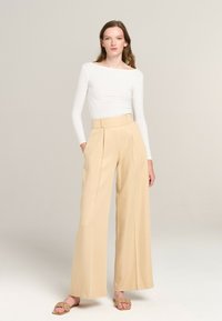 White long-sleeve top with a boat neckline, paired with high-waisted beige wide-leg trousers. Model wearing woven beige slides.