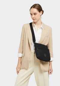 Sort crossover taske med struktureret mønster, der har lynlåslukning og et emblemer. Båret over en krämfarvet blazer og hvid skjorte.