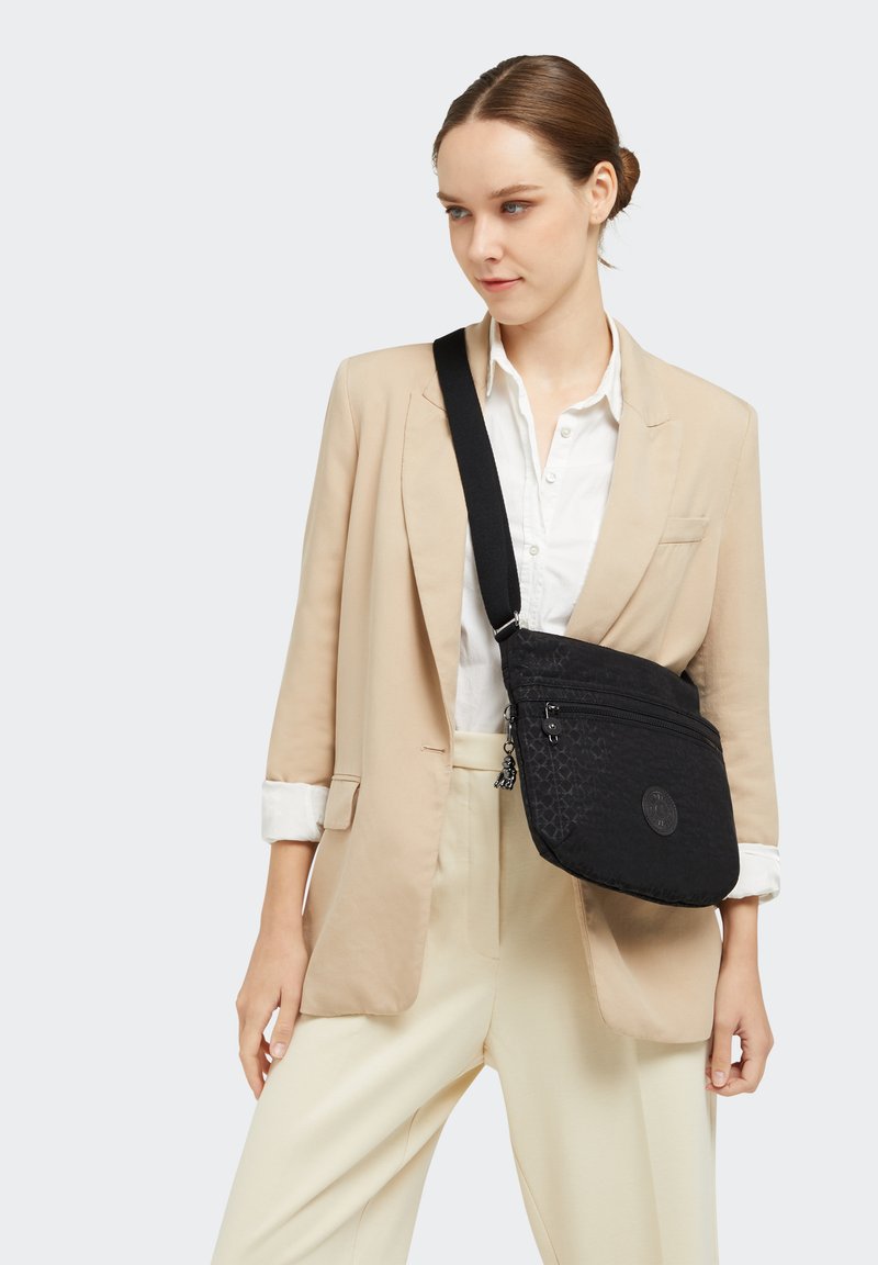 Sort crossover taske med struktureret mønster, der har lynlåslukning og et emblemer. Båret over en krämfarvet blazer og hvid skjorte.