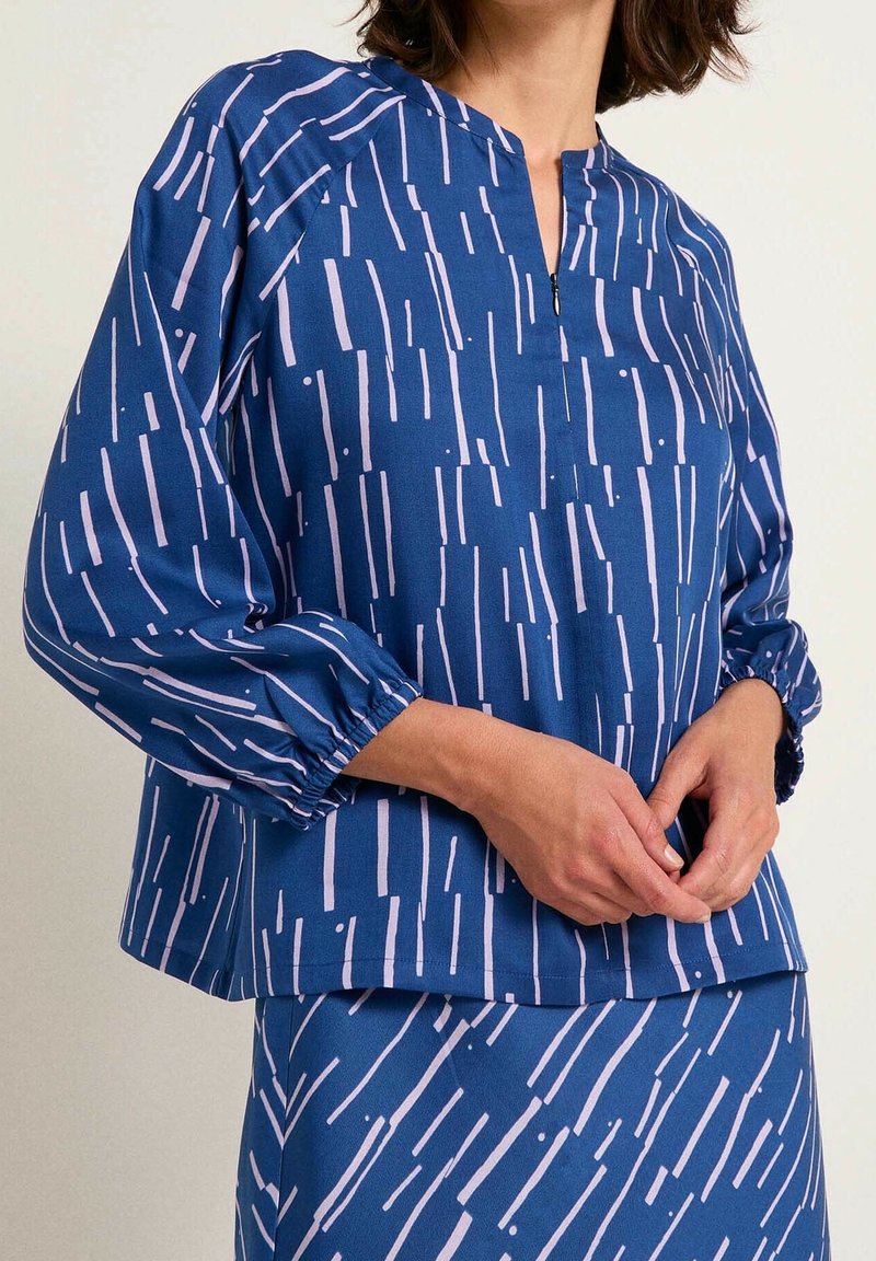 Femme portant un haut bleu à manches longues et une jupe assortie avec des motifs de lignes verticales rose clair, mains légèrement jointes devant.