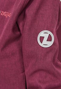 Gros plan sur la manche d'une veste bordeaux avec un patch circulaire cousu présentant un logo stylisé en forme de "Z" sur le haut du bras.