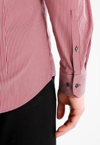 Chemise rayée rouge et blanche avec un poignet à boutons, texture lisse et ourlet arrondi, accompagnée d'un pantalon noir.