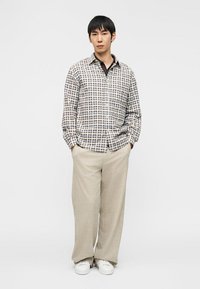 Jeune homme debout les mains dans les poches, portant une chemise à carreaux, un pantalon large beige et des baskets blanches sur un fond blanc.