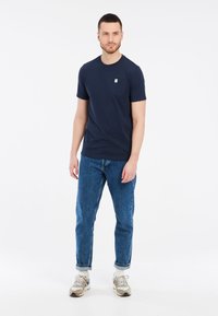 Námornícke bavlnené tričko s okrúhlym výstrihom a malou náprsní kapsou, spárované s svetlomodrými džínsami. Model má na sebe neutrálne tenisky.