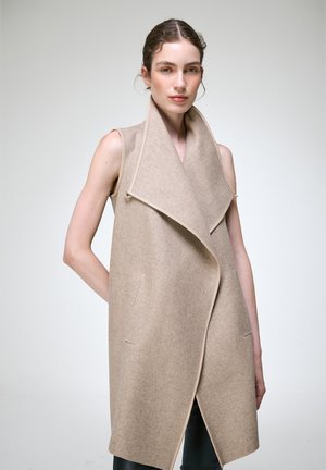 Femme portant un gilet sans manches en laine beige avec un grand col, debout devant un fond clair uni.