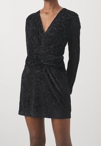Černé sametové šaty s dlouhými rukávy, hlubokým výstřihem do V a překládaným detailem v pase; mají jemný květinový vzor a hladkou texturu.