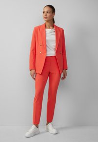 Korallenfarbener doppelt geknöpfter Blazer und dazu passende maßgeschneiderte Hose, kombiniert mit einem weißen drapierten Top und weißen Sneakern. Glatte Stofftextur.