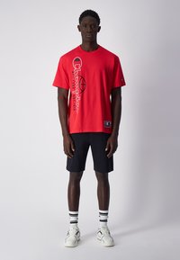 Camiseta de algodón roja con gráficos negros, cuello redondo y mangas cortas. Combinada con pantalones cortos negros y zapatillas blancas con calcetines rayados.