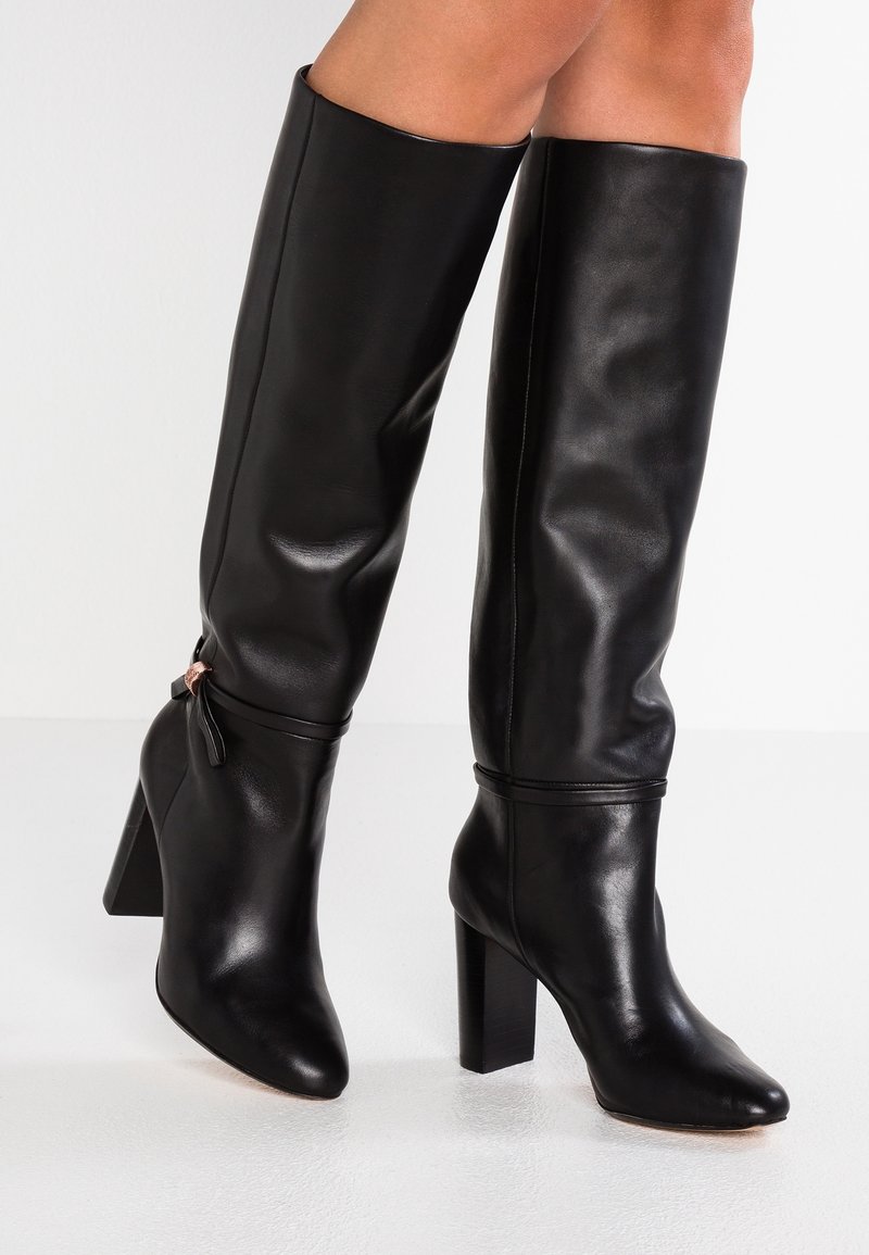 Black leather knee-high boots with a pointed toe, chunky block heel, ruching detail at the top, and a small decorative strap with hardware.