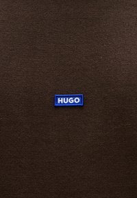 Fond en tissu marron avec une étiquette rectangulaire bleue portant l'inscription "HUGO" en lettres blanches, mettant en valeur un motif de tissage texturé.