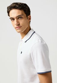 Weißes Polo-Shirt aus strukturiertem Stoff, mit zwei Knöpfen, einem Kragen und Ärmelabschlüssen in Marineblau gestreift. Kleines Emblem auf der Brust.