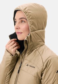 Beige geïsoleerde jas met een capuchon, voorzien van een zwarte binnenvoering, ritssluiting en een gequiltde textuur. Logo weergegeven op de linkerborst.