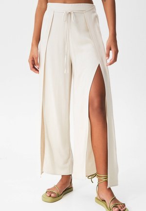 Beige wijde broekspijpen met hoge zijopeningen en een taille met trekkoord, gedragen met olijfgroene platte sandalen met bandjes, gedragen door een staand persoon.