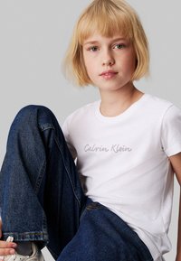 Camiseta de algodón blanca con logo "Calvin Klein" en gris claro, combinada con jeans de denim azul oscuro, con un ajuste relajado y puños ligeramente doblados.