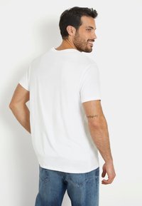Tricou alb din bumbac, cu guler rotund și mâneci scurte, cu o textură netedă și tiv drept, purtat cu blugi.