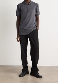 Polo gris avec deux boutons, manches courtes et logo subtil. Pantalons noirs à coupe droite et texture solide. Chaussures noires.