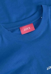Modrá bavlněná tričko s kulatým výstřihem a hladkým povrchem. Na přední straně se nachází růžová nášivka s nápisem "JJXX" a malé bílé logo.