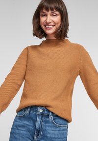 Bruine geribde trui met een hoge hals en lange mouwen, gecombineerd met blauwe denim jeans. Gestructureerde gebreide stof met een losse pasvorm.